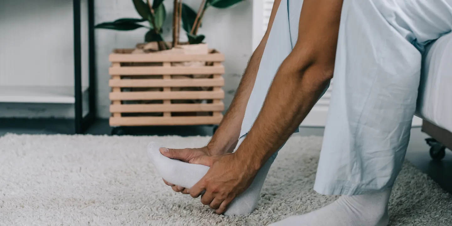 Man sitting on the edge of a bed pressing into his heel and arch during painful first steps of the morning