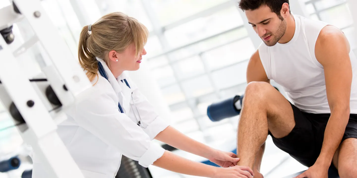 Physiotherapist checking a seated athlete’s ankle in a gym part-way through recovery from a sprain