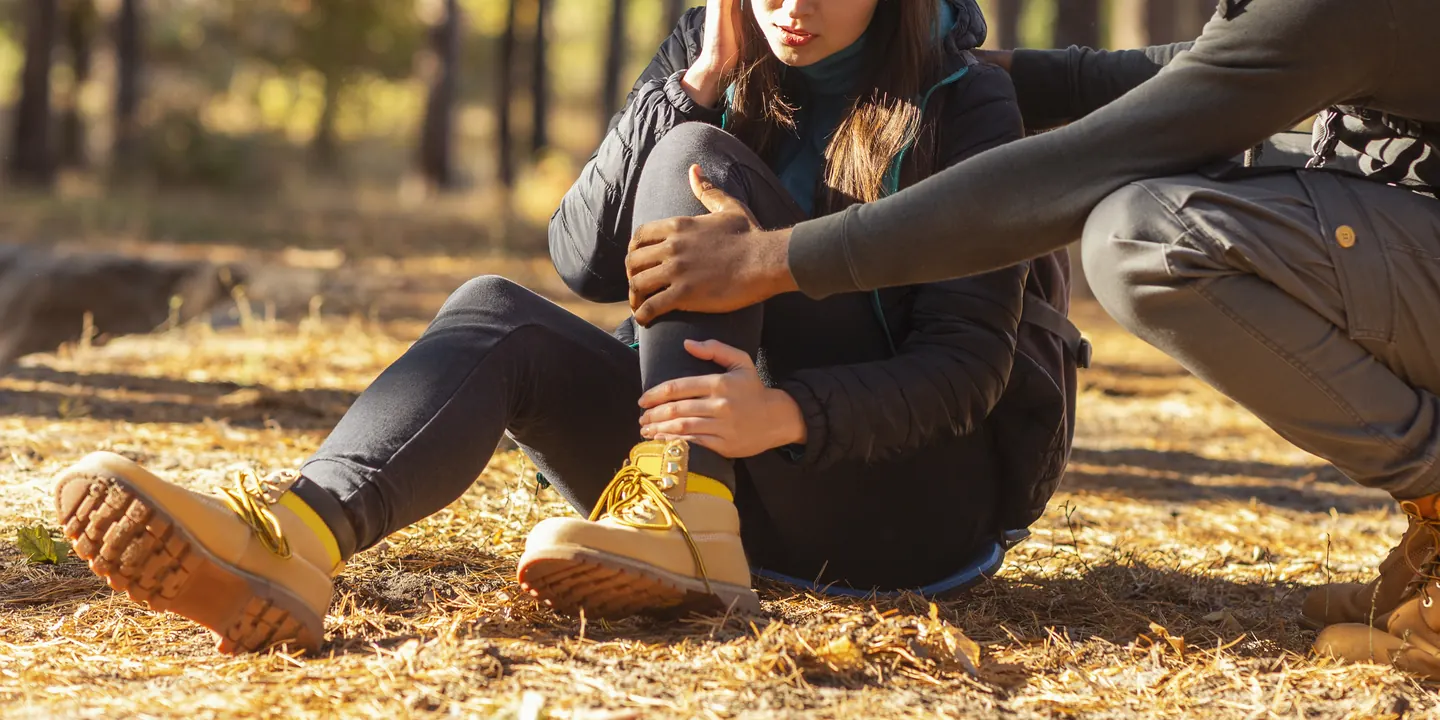Person sitting on the ground outdoors holding an injured ankle while a companion crouches to help