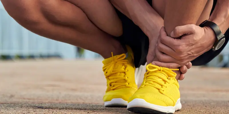 Runner in yellow trainers crouching on a path, gripping the back of the ankle where the Achilles tendon hurts